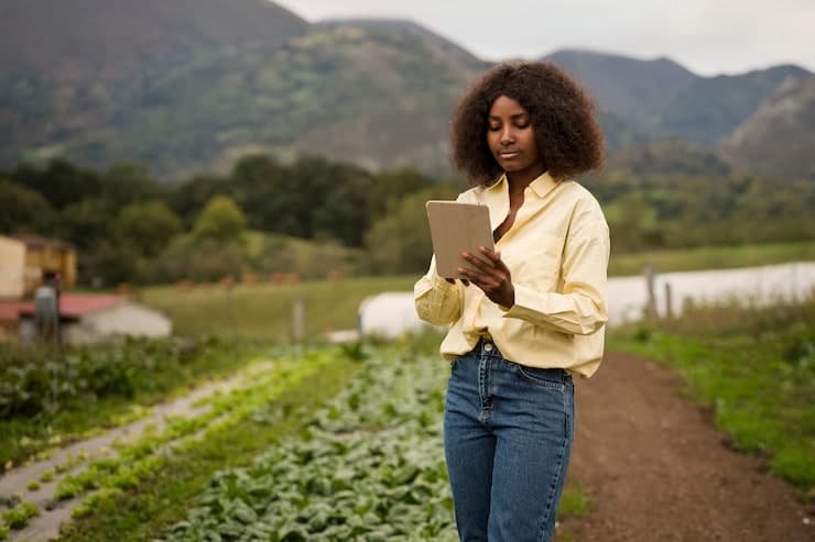 A farmer reading a tablet in a field with hills behind her